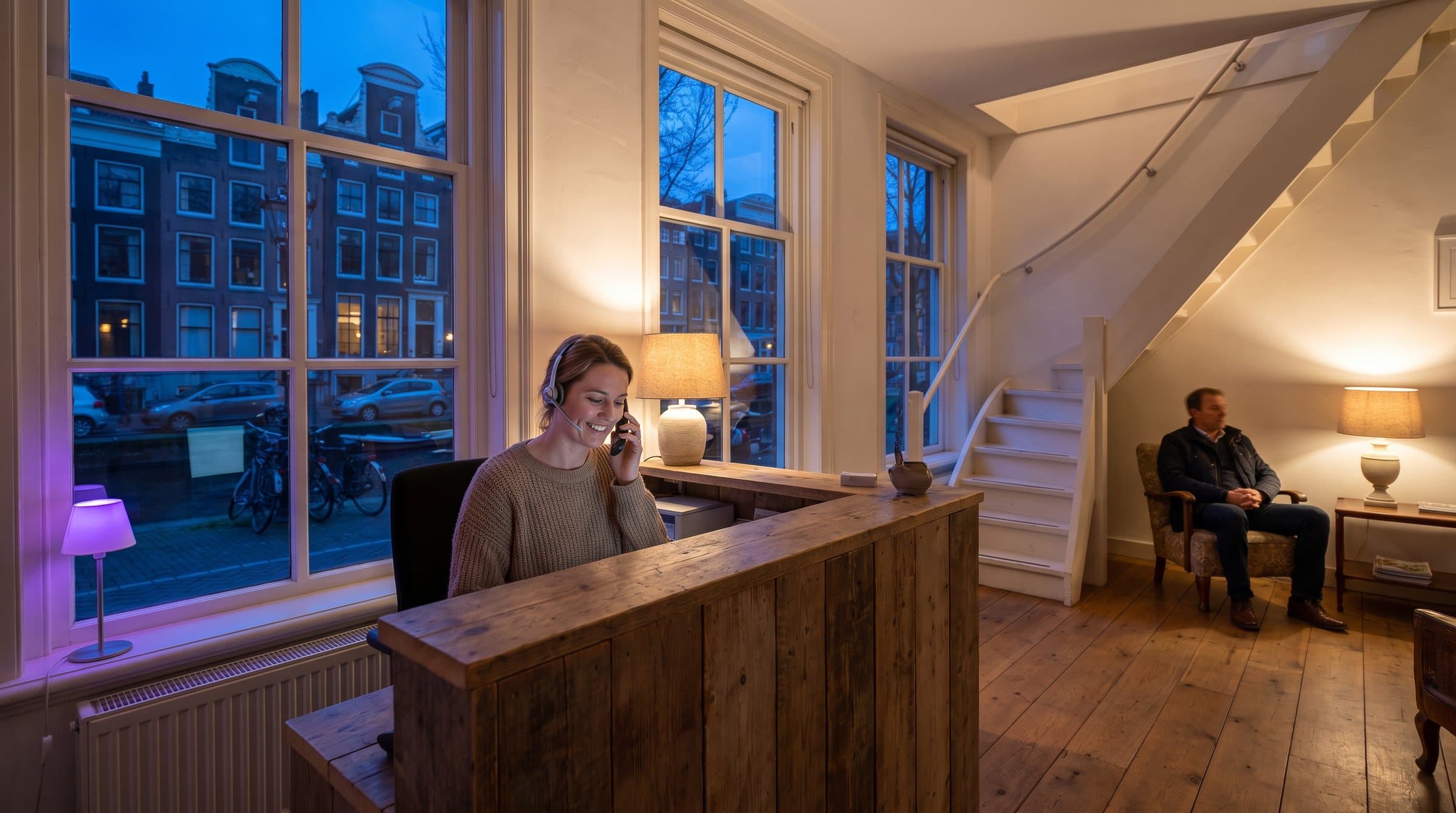 Receptioniste met headset aan de telefoon bij de balie van een tandartspraktijk in een Amsterdams grachtenpand, met uitzicht op de gracht tijdens het blauwe uur